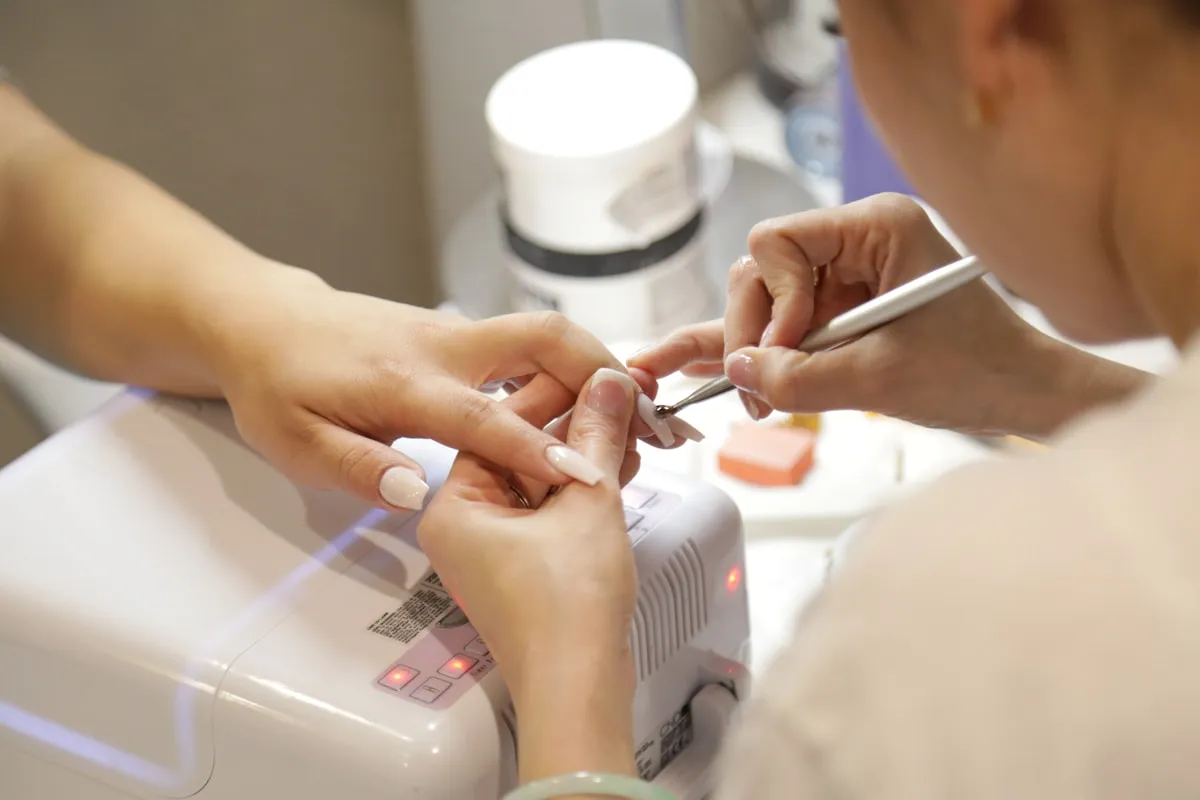 Mãos de manicure trabalhando na unha de uma cliente, com os potes de esmalte sobre a mesa